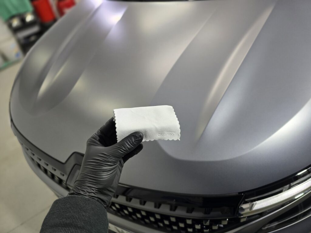 Mano con guante negro limpiando capó de coche gris.